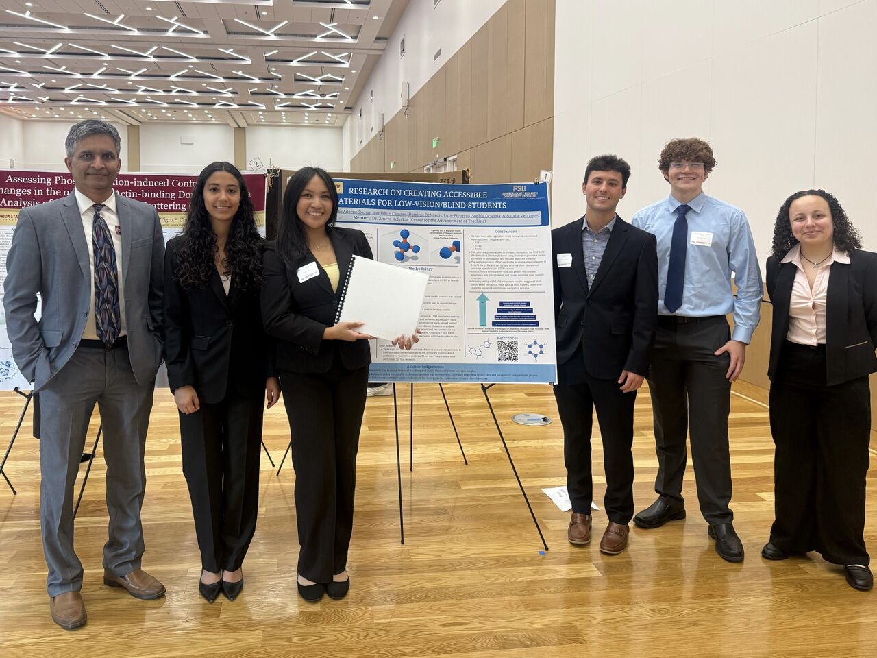 5 of 6 research students presenting at the poster session. They are standing in front of the poster with their mentor, Dr. Kolarkar, standing at the far left. One student is holding the braille booklet of the poster.