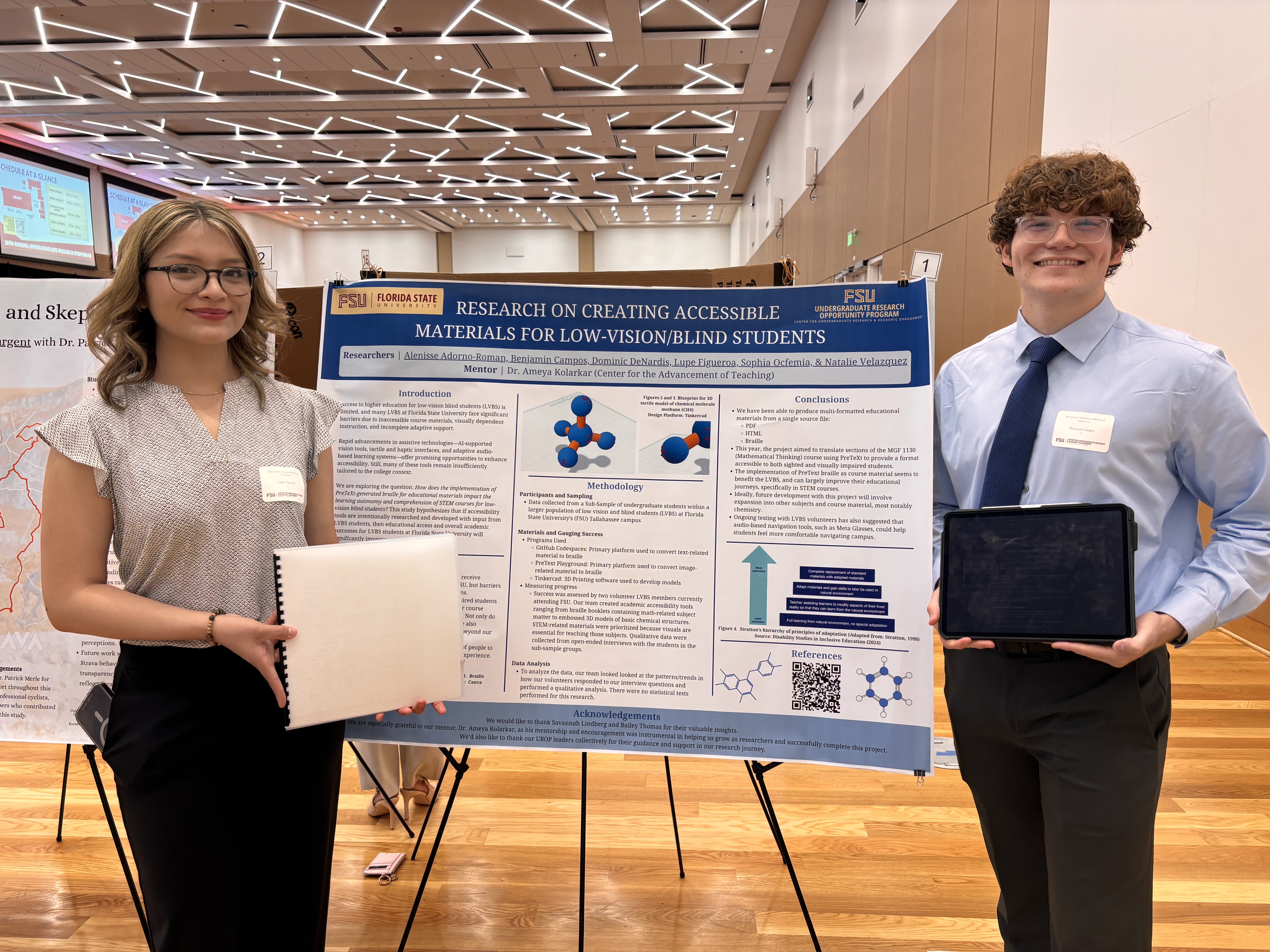 2 research students are standing in front of the poster. One student is holding the braille booklet of the poster.